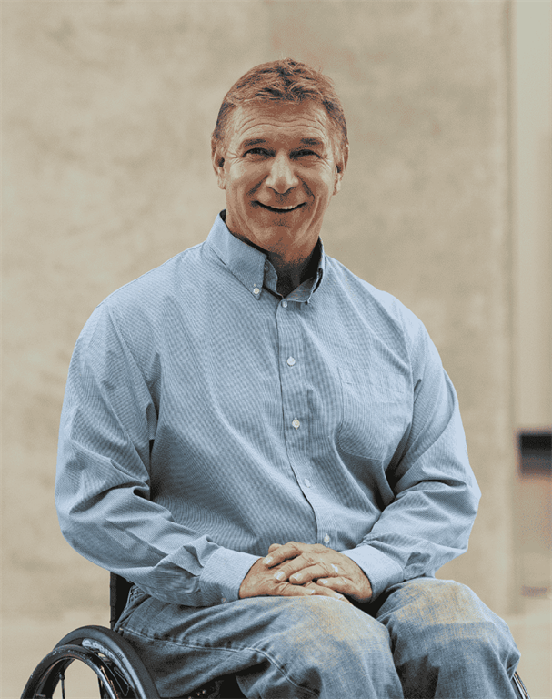 Rick Hansen smiles for a professional photo. He is in a blue button shirt and blue jeans.